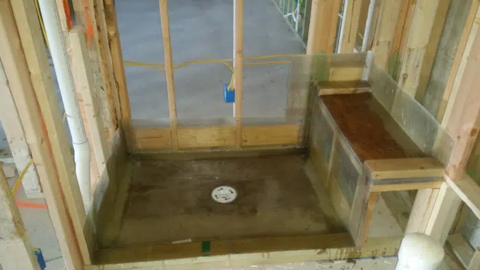 Membrane-coated shower pan sloped to a capped drain, with framed corner bench at right