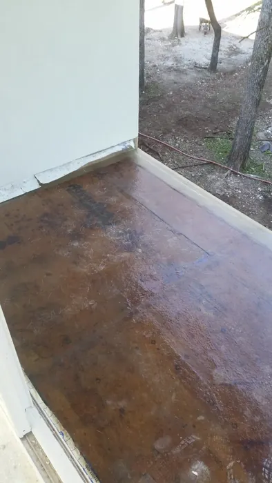 Exterior deck coated in brown waterproofing resin, bounded by light grey walls above bare yard