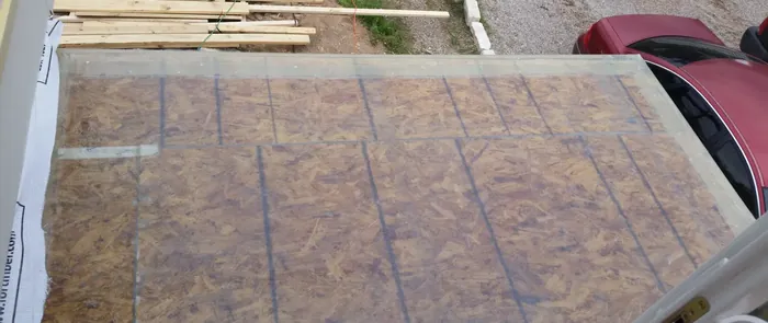 Exterior deck platform sheathed in OSB with chalked layout grid, lumber stacked on the ground beyond