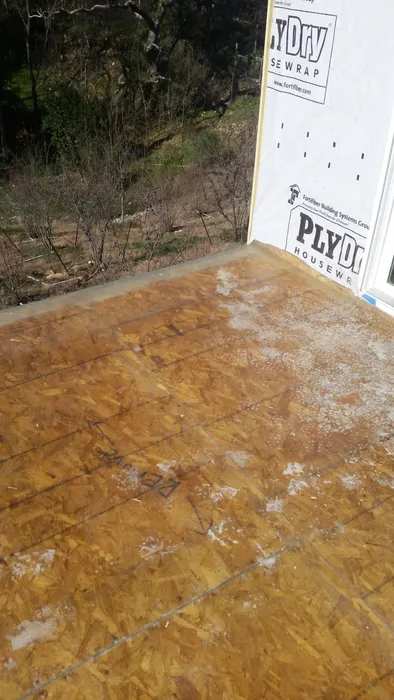 Bare OSB deck sheathing meeting a housewrapped wall, wooded slope beyond, surface prepped before coating